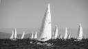 Black and white photo of a fleet of small sailboats racing on open sea, with one boat, sail number 3102, in front and hills in the background.
